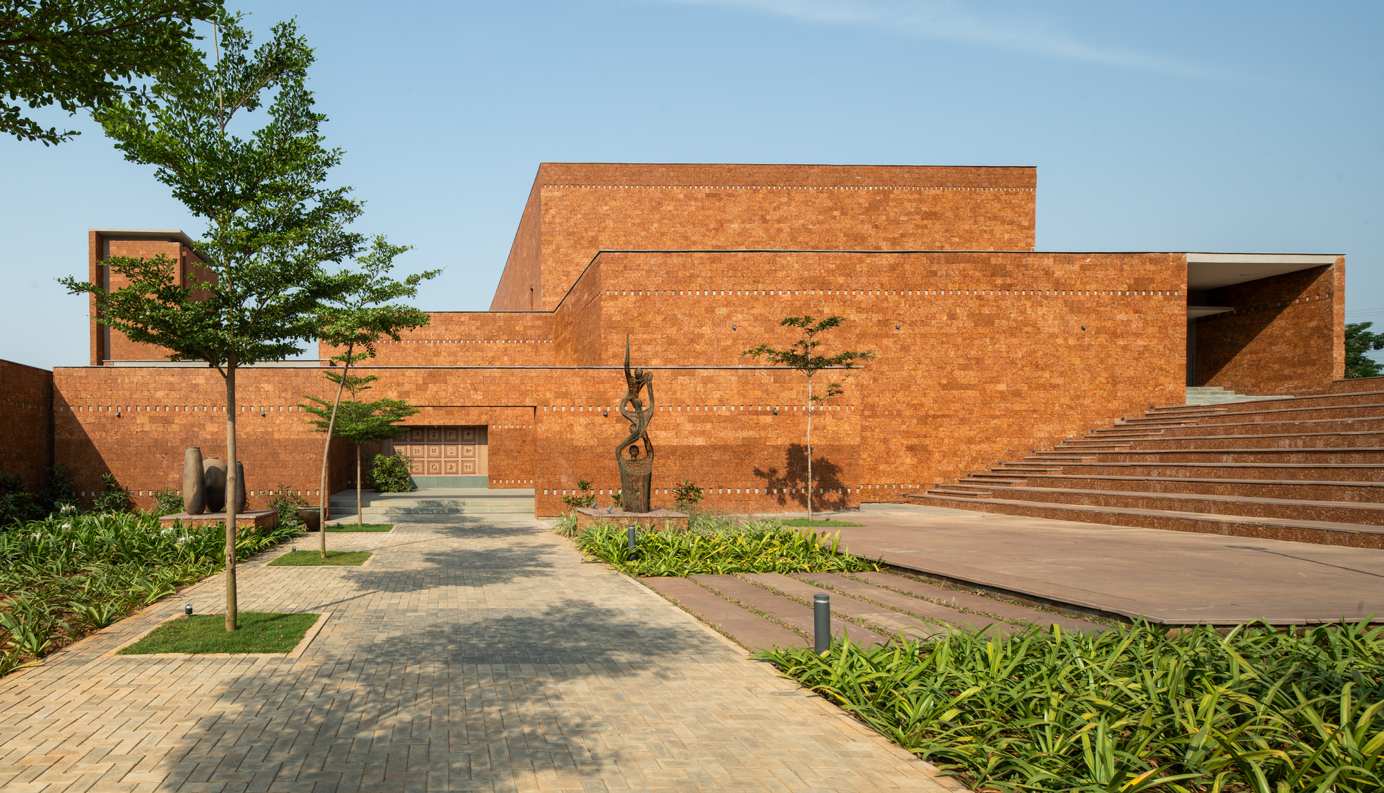 Red laterite brick auditorium volume rising behind a plaza with a small bronze sculpture