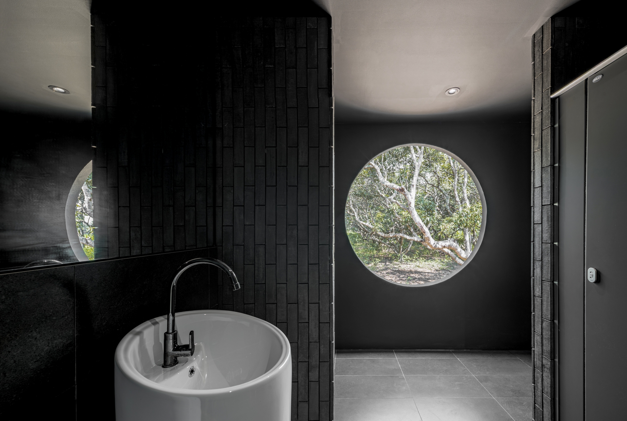 Dark-tiled bathroom with a round picture window framing the mango trees