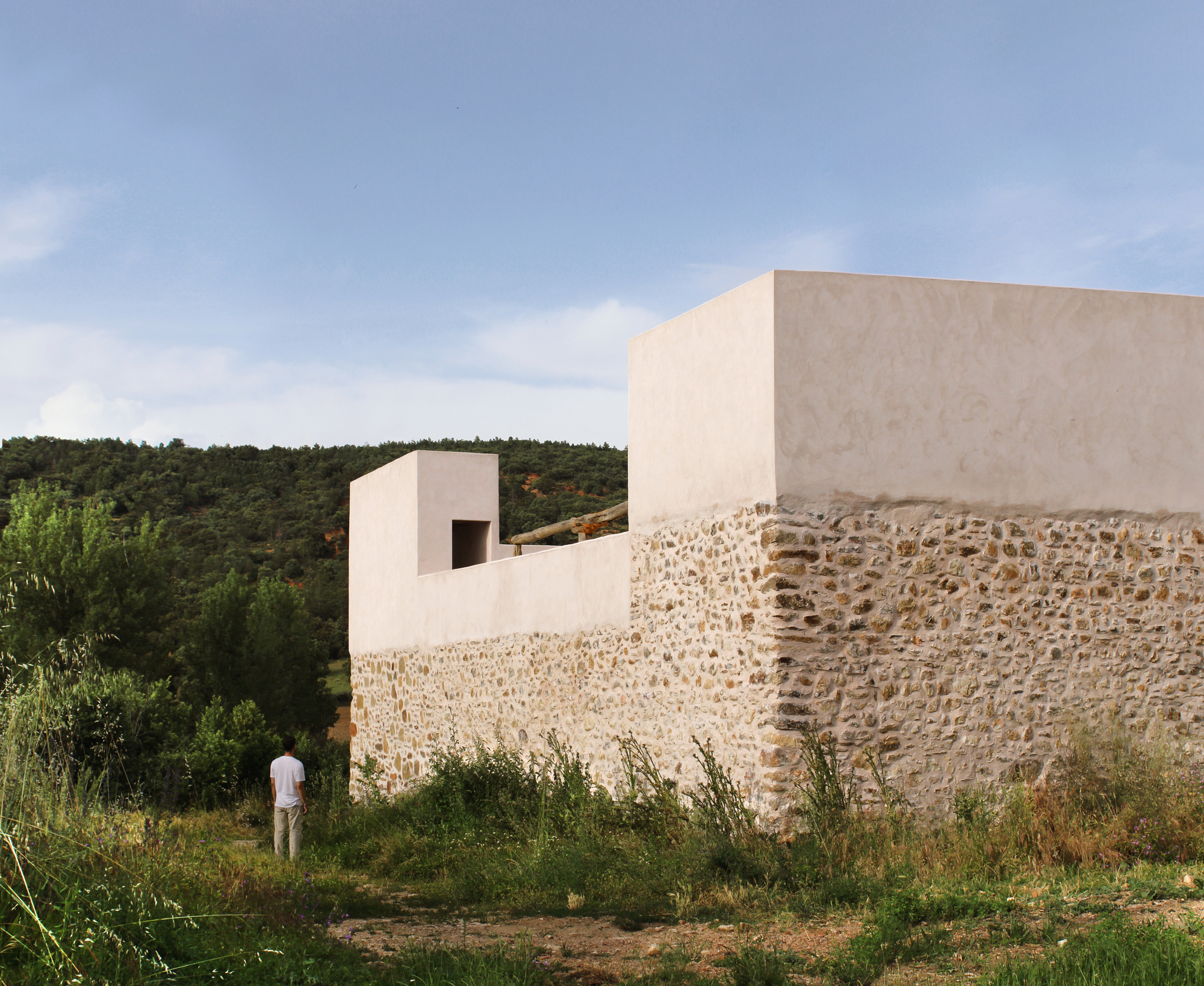 Long view of the white plaster volume meeting the rough stone perimeter wall with a figure standing in the grass