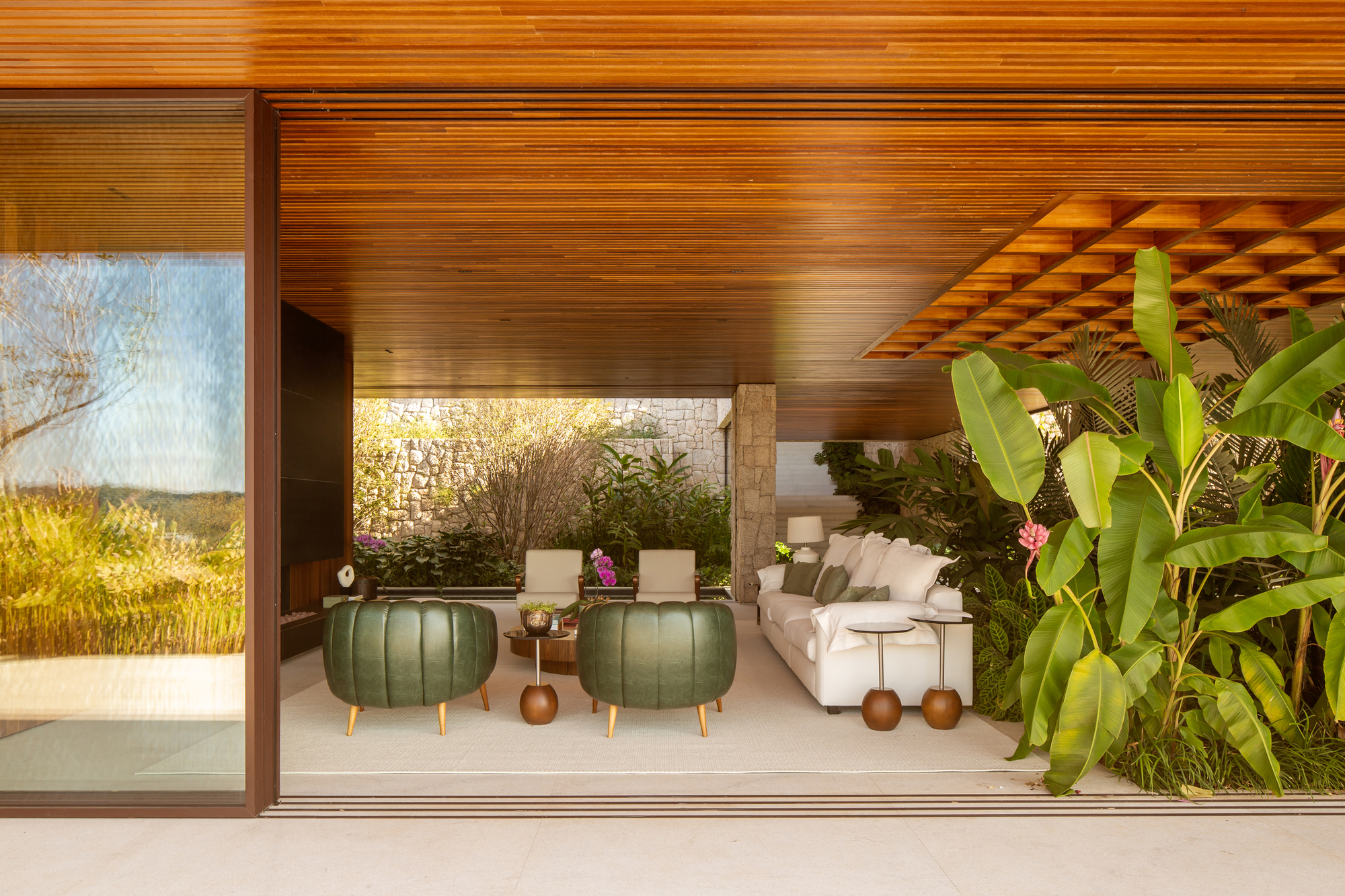 Living lounge with green leather pouffes, a low timber coffered canopy and glass walls opening to the garden